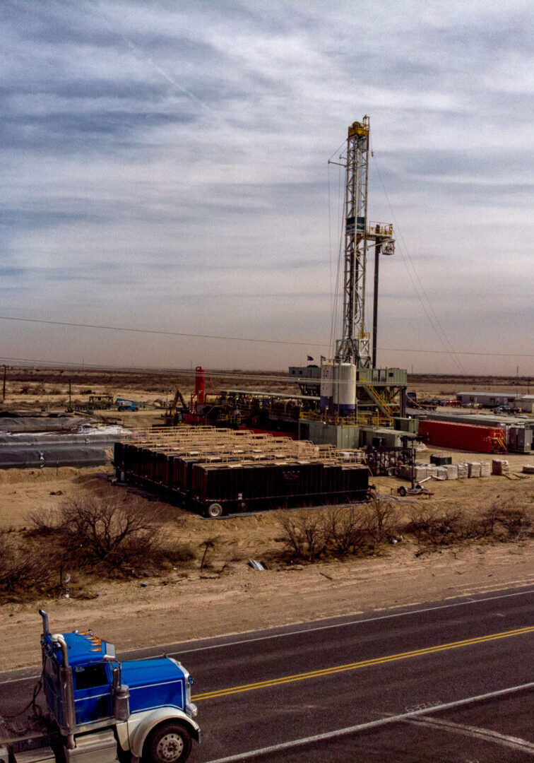 A Semi Truck Hauling Frack Water Drives Past A Drilling Rig Site That Works To Pull Crude Oil And Gas Out Of The Ground To Be Refined